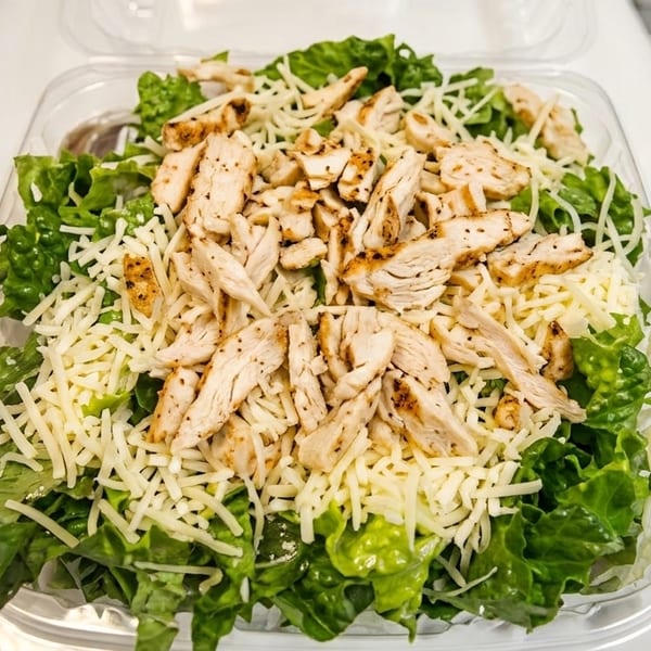 Grilled chicken and shredded cheese on a bed of fresh green lettuce in a clear plastic container.